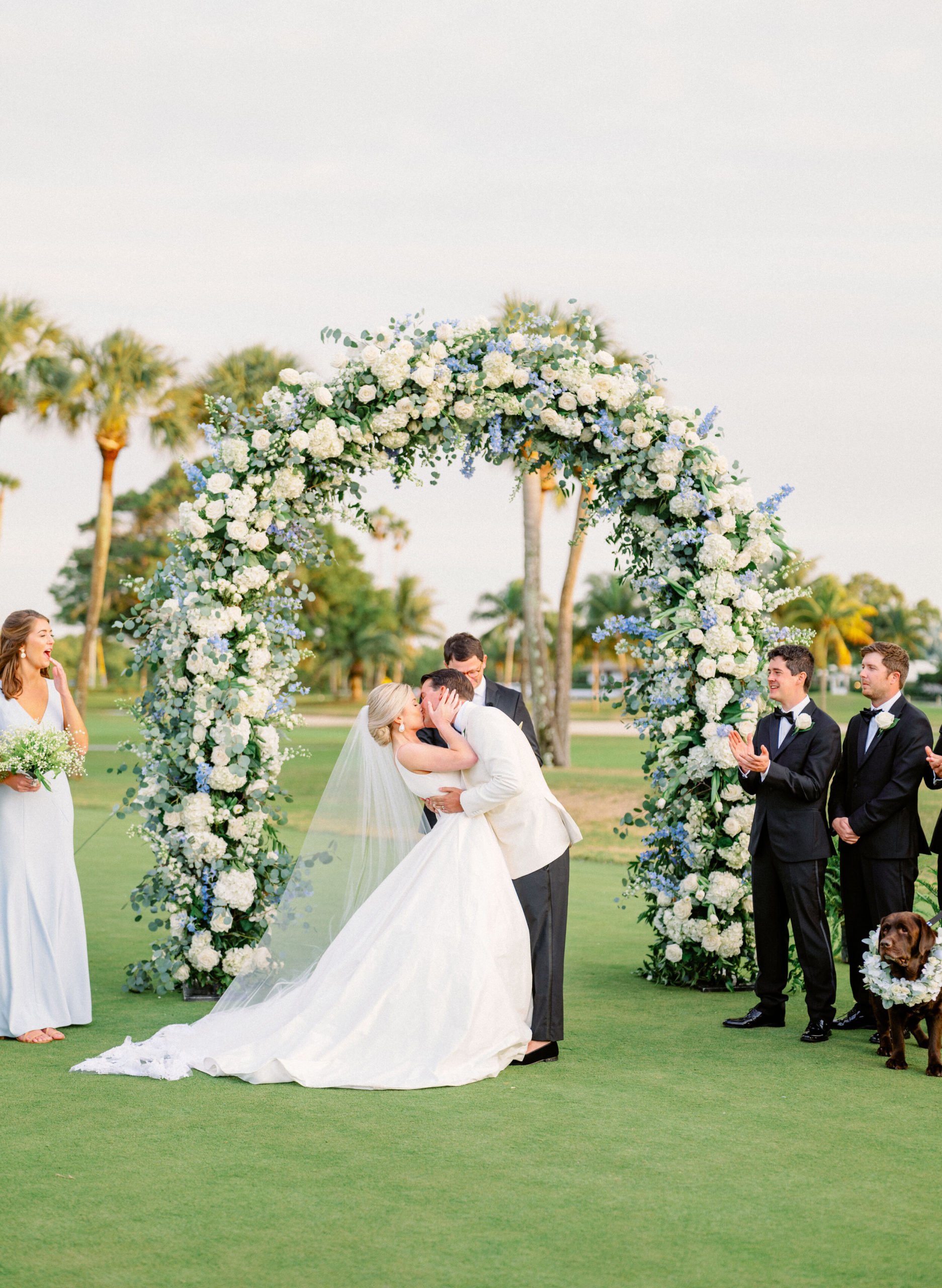 couple kissing beneath floral arch of white and blue flowers at Gasparilla Inn outdoor ceremony