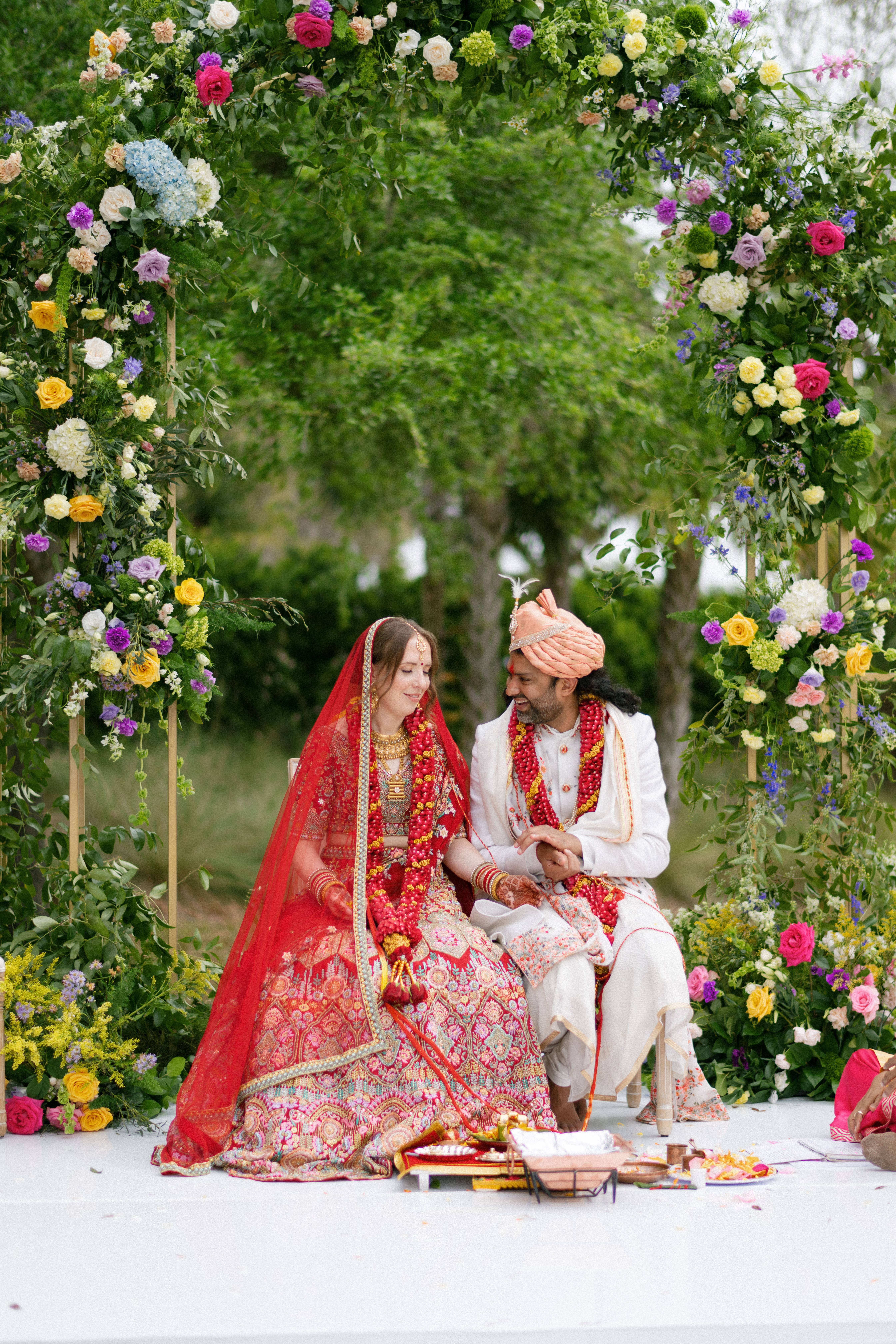 A bride and groom in traditional Indian attire seated under a floral arch.