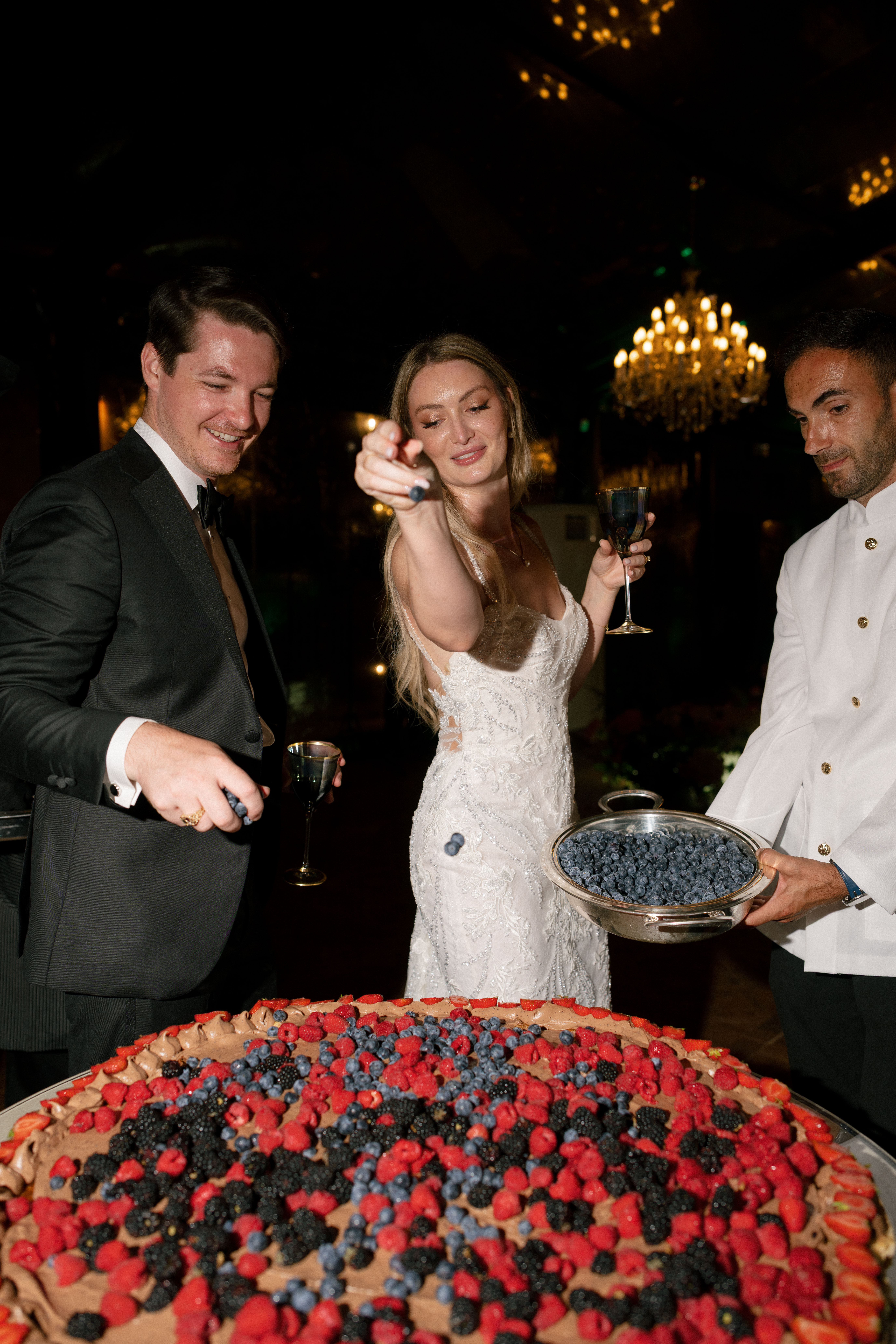 Bride and groom decorating an Italian wedding cake with berries and powdered sugar.