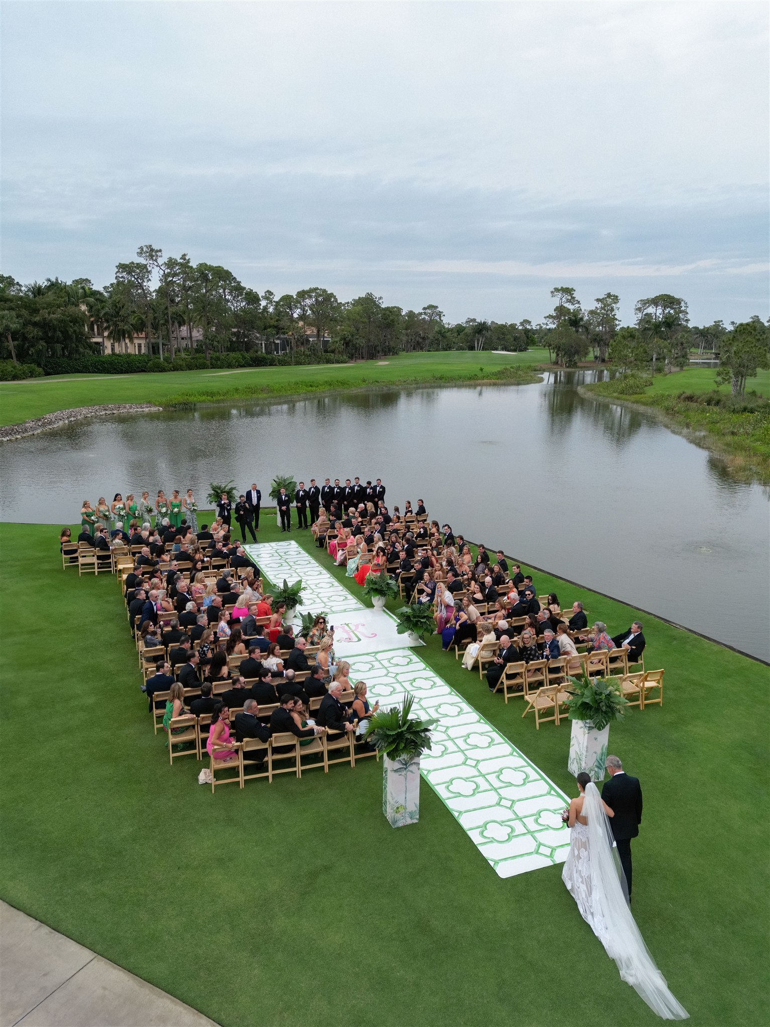 Aerial shot of an outdoor wedding by the water with a custom aisle atop the lawn.