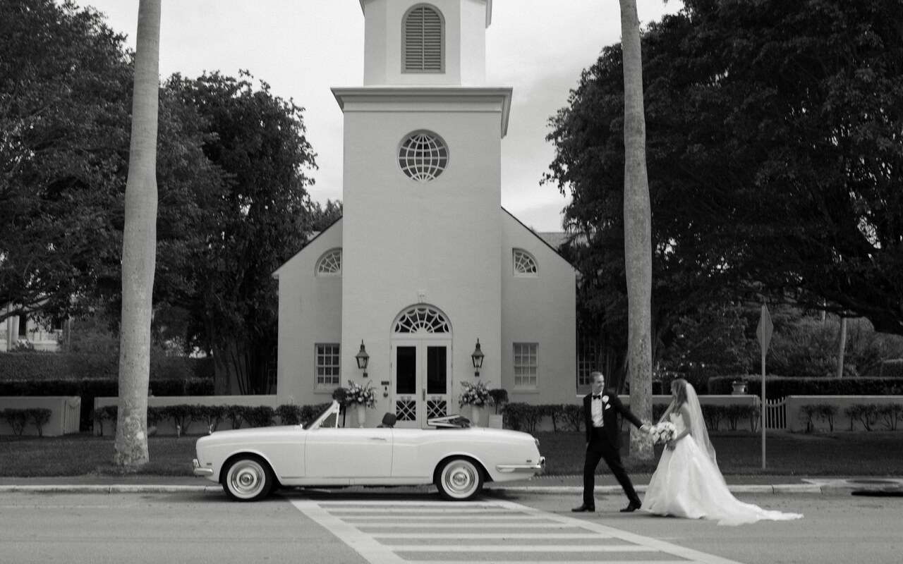 Black and white portrait of a bride and groom outside of their wedding church ceremony with a classic car.