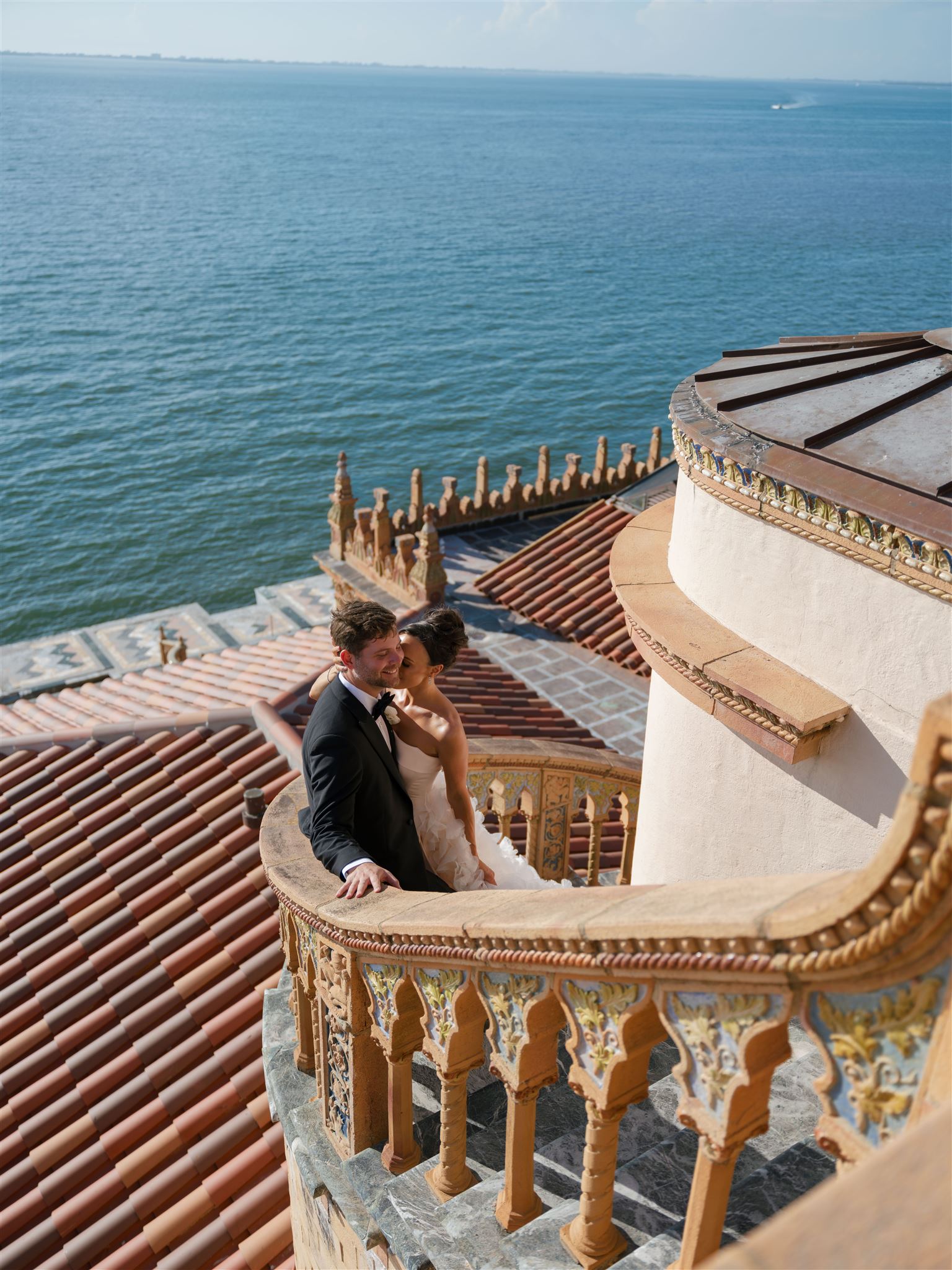 A bride and groom posing for portraits on the stairs of The Ringling in Florida, the blue ocean sprawling behind them.