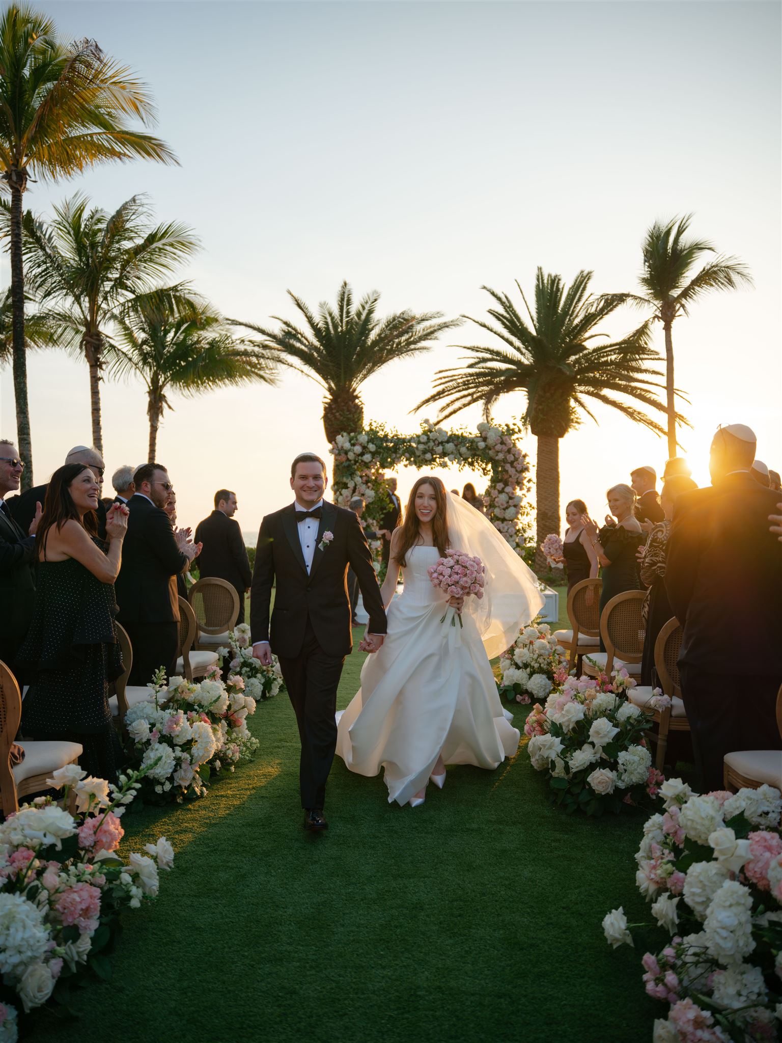 A bride and groom walk back down the aisle surrounded by palm trees and blush and cream florals.