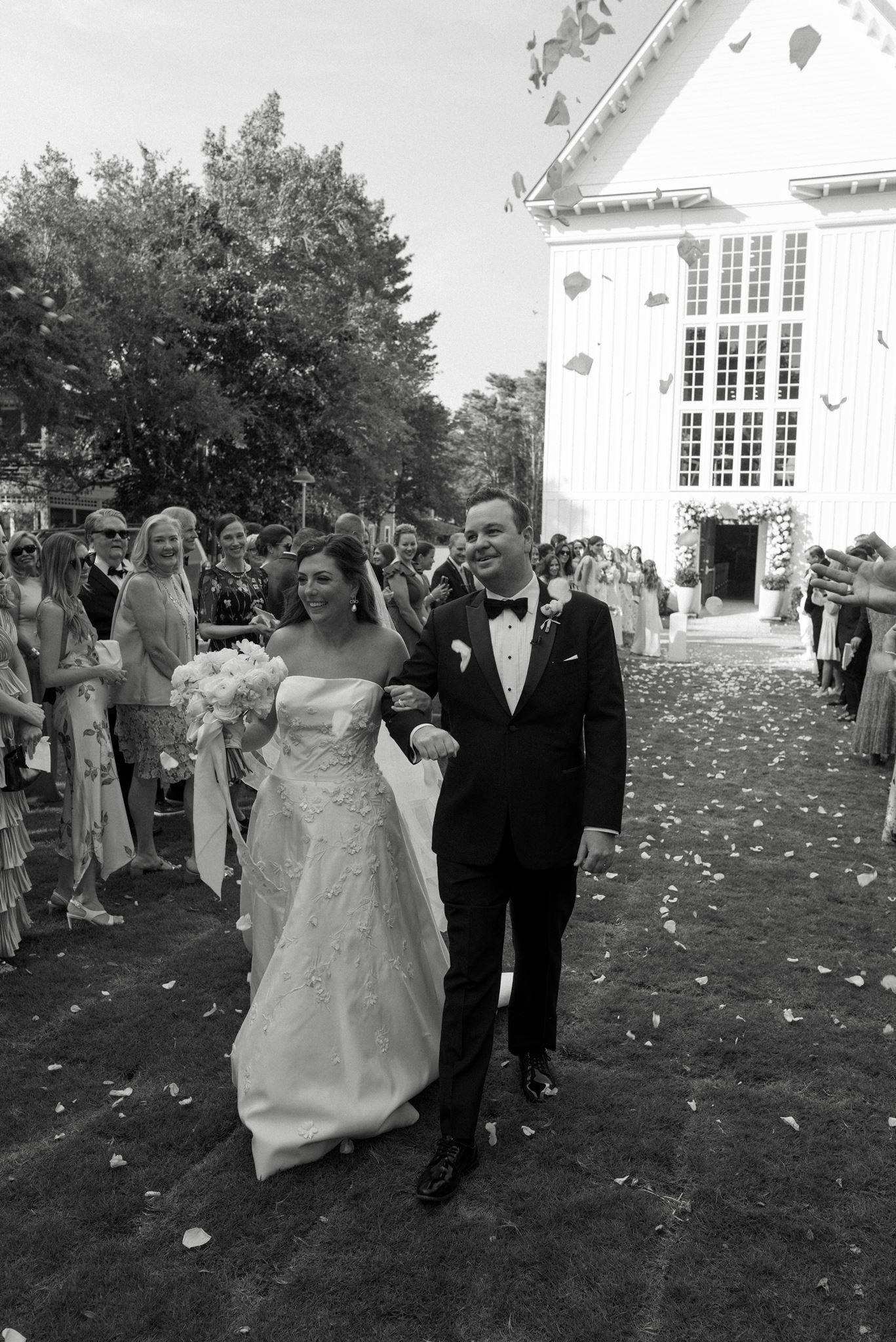 A bride and groom walking out of their church chapel ceremony surrounded by guests and flying flower petals.