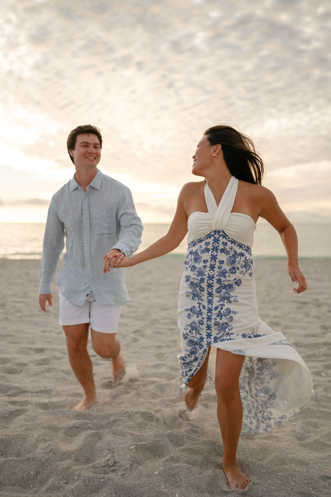 An engaged couple runs down the beach at golden hour, hand in hand.