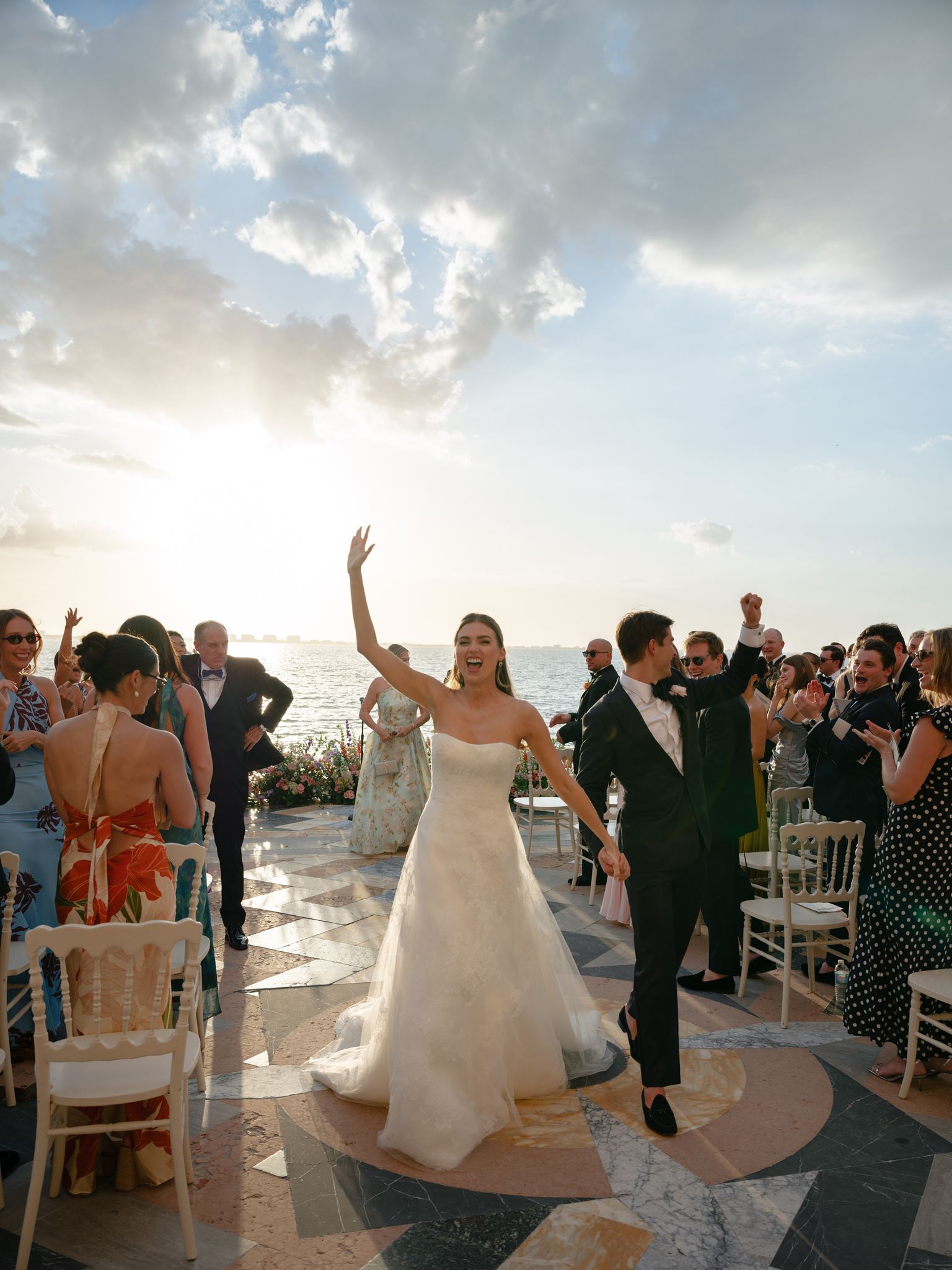 A bride and groom coming back down their outdoor aisle at sunset, golden sun and sparkling ocean behind them.