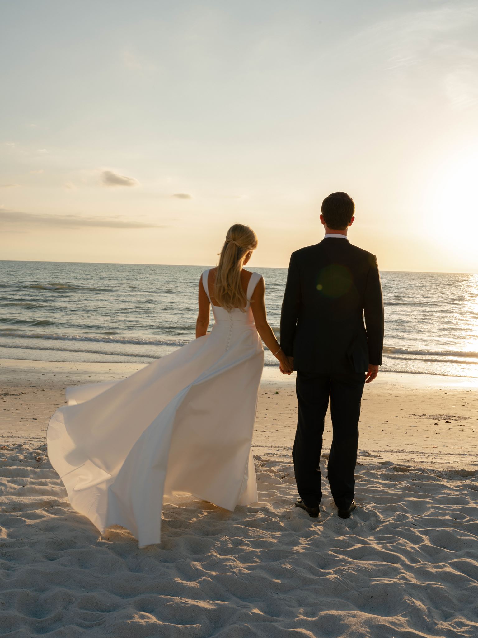 A bride and groom stand on a Florida beach hand in hand as the sun sets over the ocean.