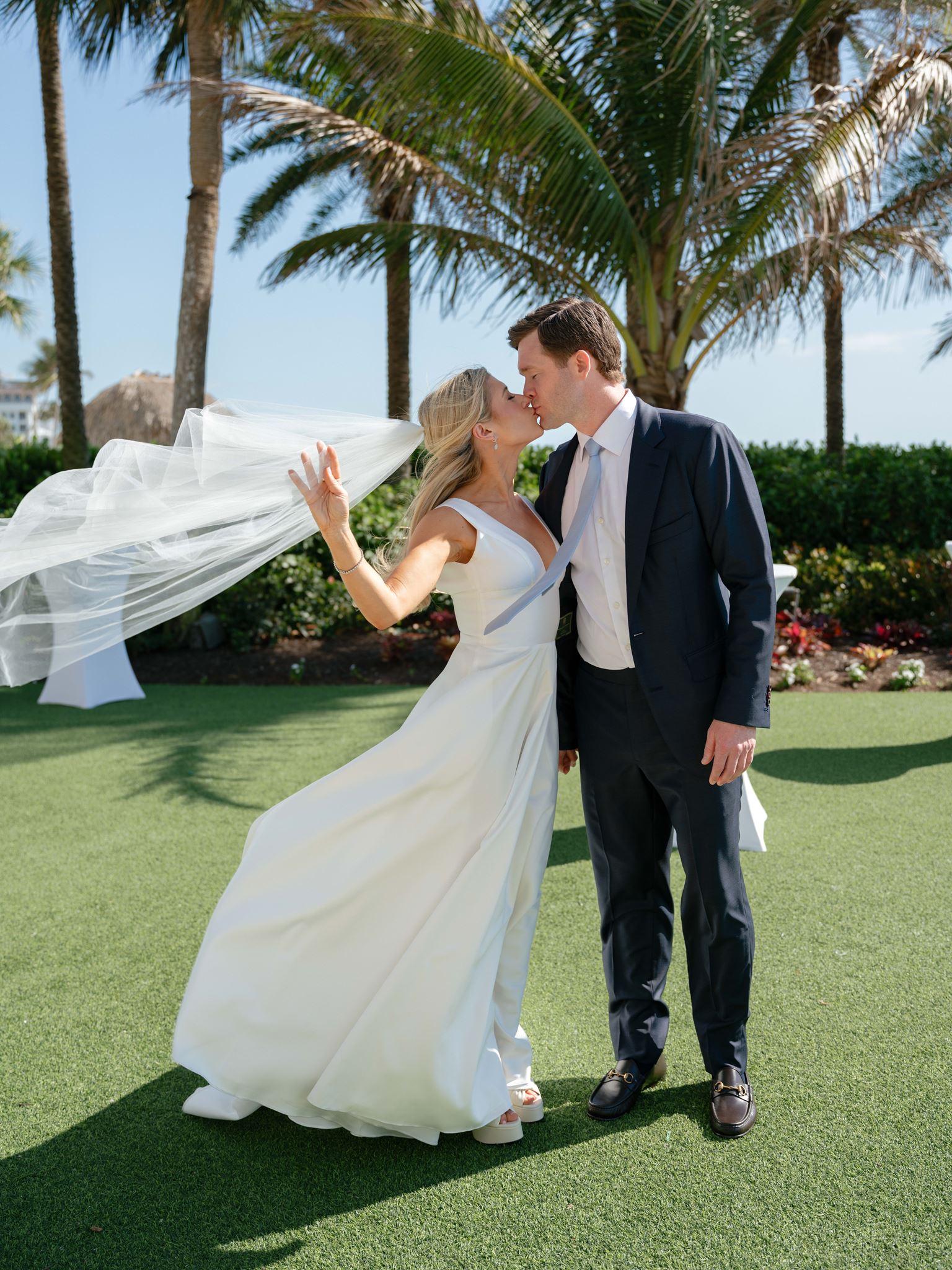 Bride and groom kissing beneath the Florida palm trees as her veil flows behind her.